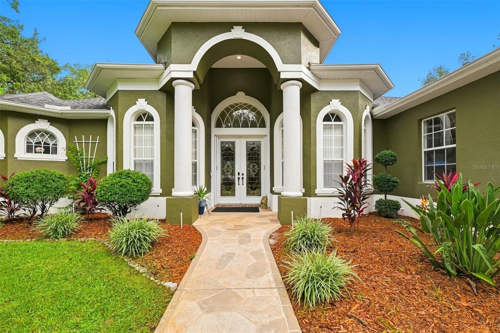 A front view of a green house with white columns, arched windows, and a stone walkway — 16547 CROSSANDRA LANE, SPRING HILL, FL