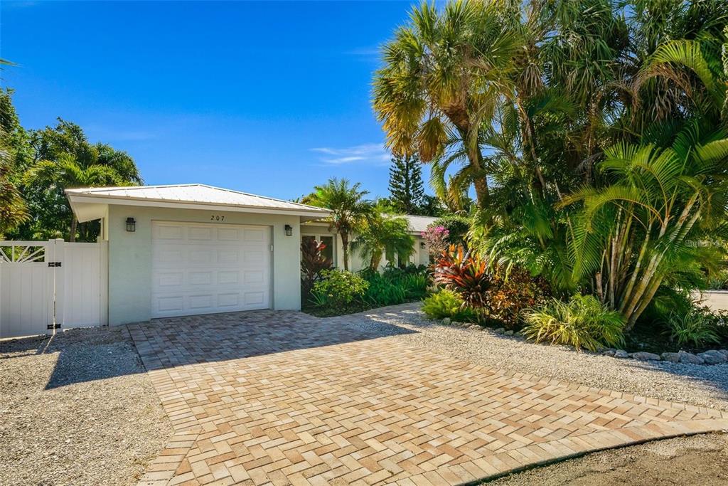 A single-story house with a white garage door, brick driveway, and lush tropical landscaping — 207 PEACOCK LANE, HOLMES BEACH, FL