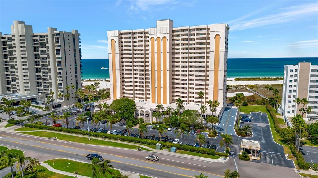 An aerial view shows a tall, multi-story residential building with a parking lot near the beach — 1270 GULF BOULEVARD UNIT 1003, CLEARWATER, FL