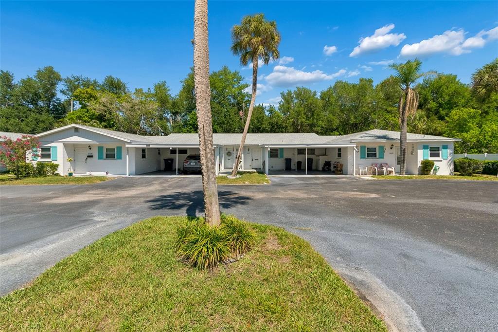 A single-story multi-unit building with white siding, teal shutters, and attached carports under a blue sky — 6072 ARLENE WAY, BRADENTON, FL