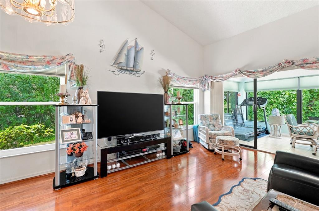 A living room features hardwood floors, a large television, glass shelving, and a sunroom beyond — 9239 TURNBERRY COURT, NEW PORT RICHEY, FL