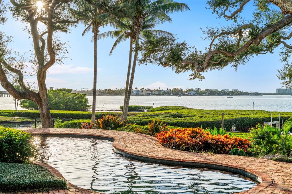 A curved brick-edged pool overlooks a bay with palm trees and a distant shoreline — 3000 SOUTHWEST DRIVE, SARASOTA, FL