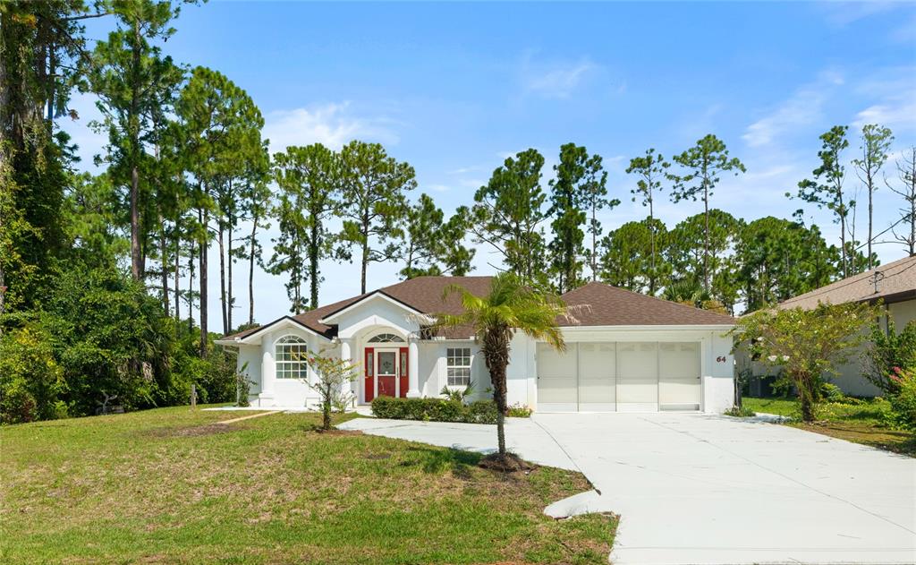 A white single-story house with a red front door, three-car garage, and surrounding pine trees — 64 WHITTINGTON DRIVE, PALM COAST, FL