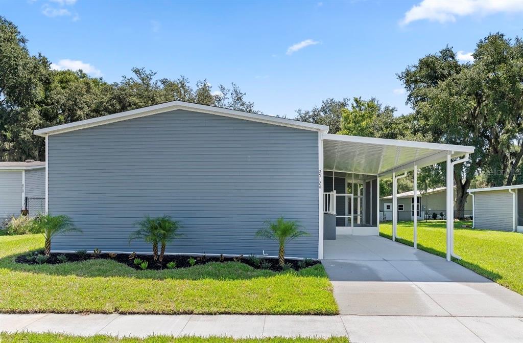 A blue-sided manufactured home features a concrete driveway, a covered carport, and small palm trees — 25124 HUCKLEBERRY WAY, ASTATULA, FL