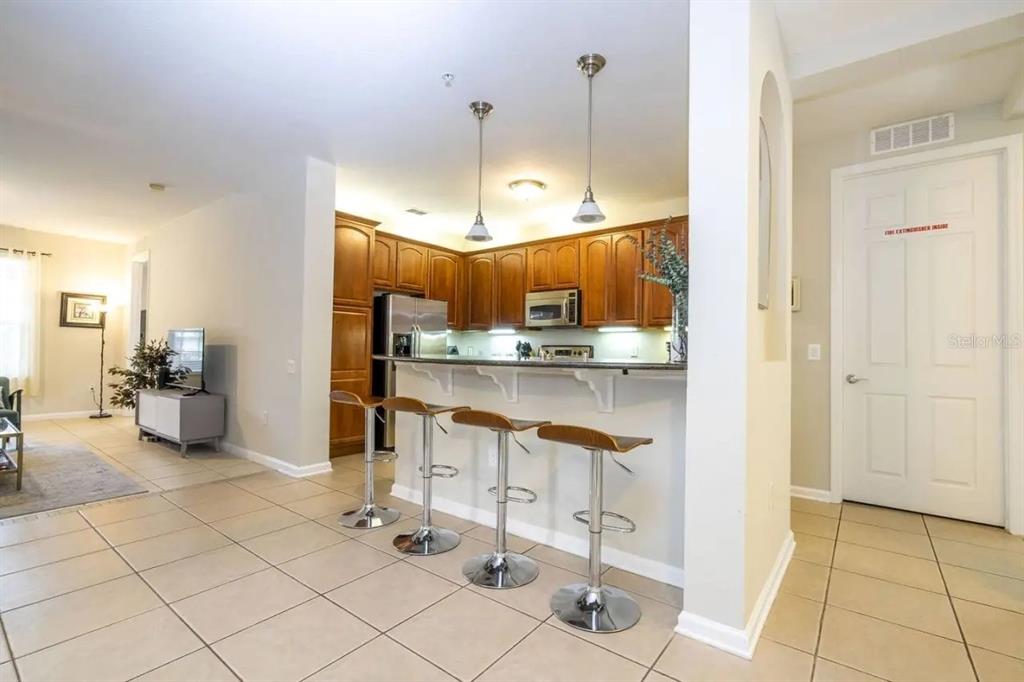 A kitchen with wood cabinets and a breakfast bar with three stools faces a living area — 5025 SHOREWAY LOOP UNIT 10703, ORLANDO, FL