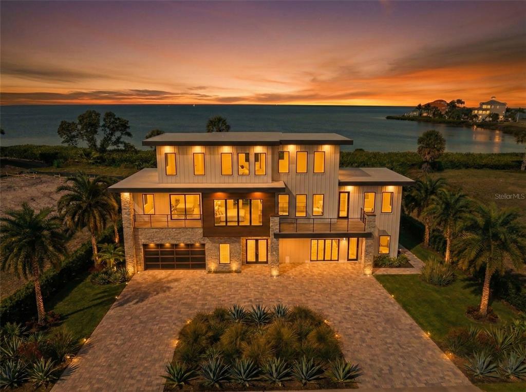 A modern three-story waterfront home with a paved driveway and palm trees at sunset — 26 HARBORPOINTE DR, PORT RICHEY, FL