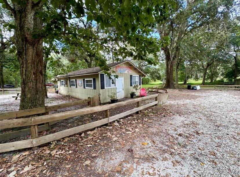 A small, light-colored building with a wooden fence sits on a gravel lot under trees — 24392 LANARK ROAD, BROOKSVILLE, FL