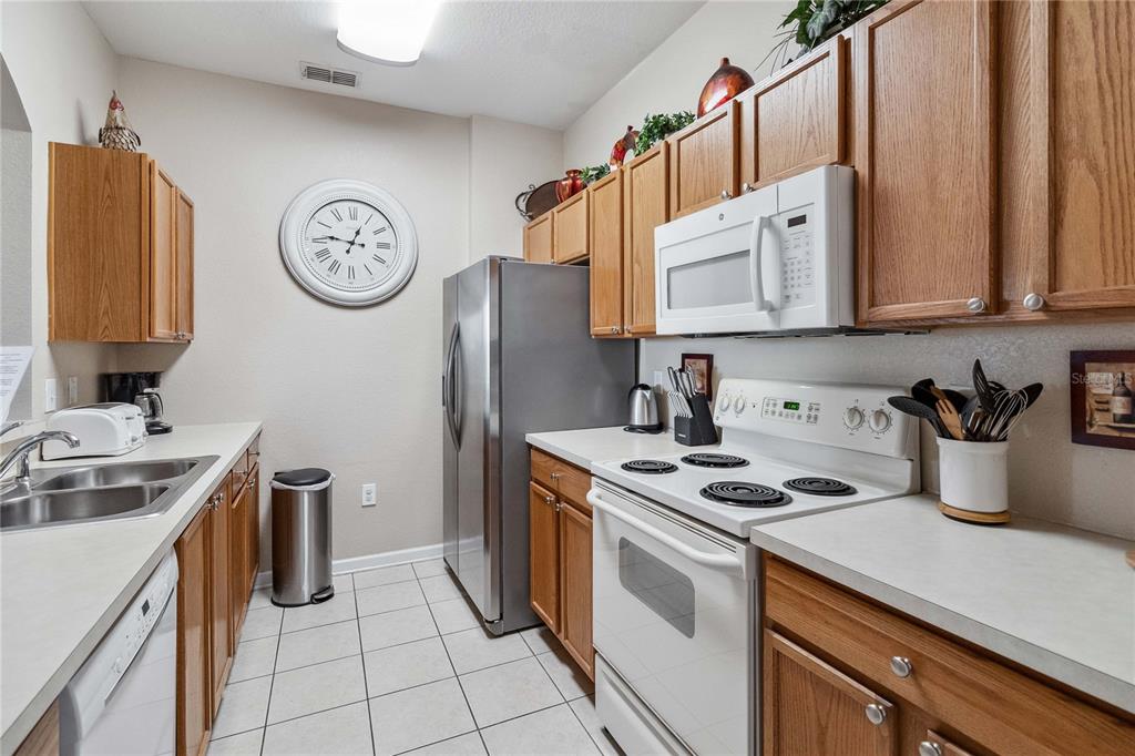 A galley kitchen features oak cabinets, white appliances, a stainless steel refrigerator, and tiled flooring — 2821 E ALMATON LOOP UNIT 401, KISSIMMEE, FL