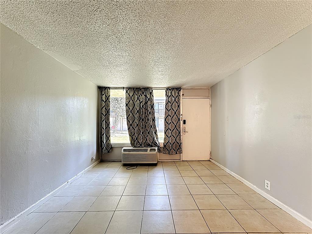 An empty room with light-colored tile flooring, gray walls, a window with curtains, and an AC unit — 7900 S ORANGE BLOSSOM TRAIL UNIT 1108, ORLANDO, FL