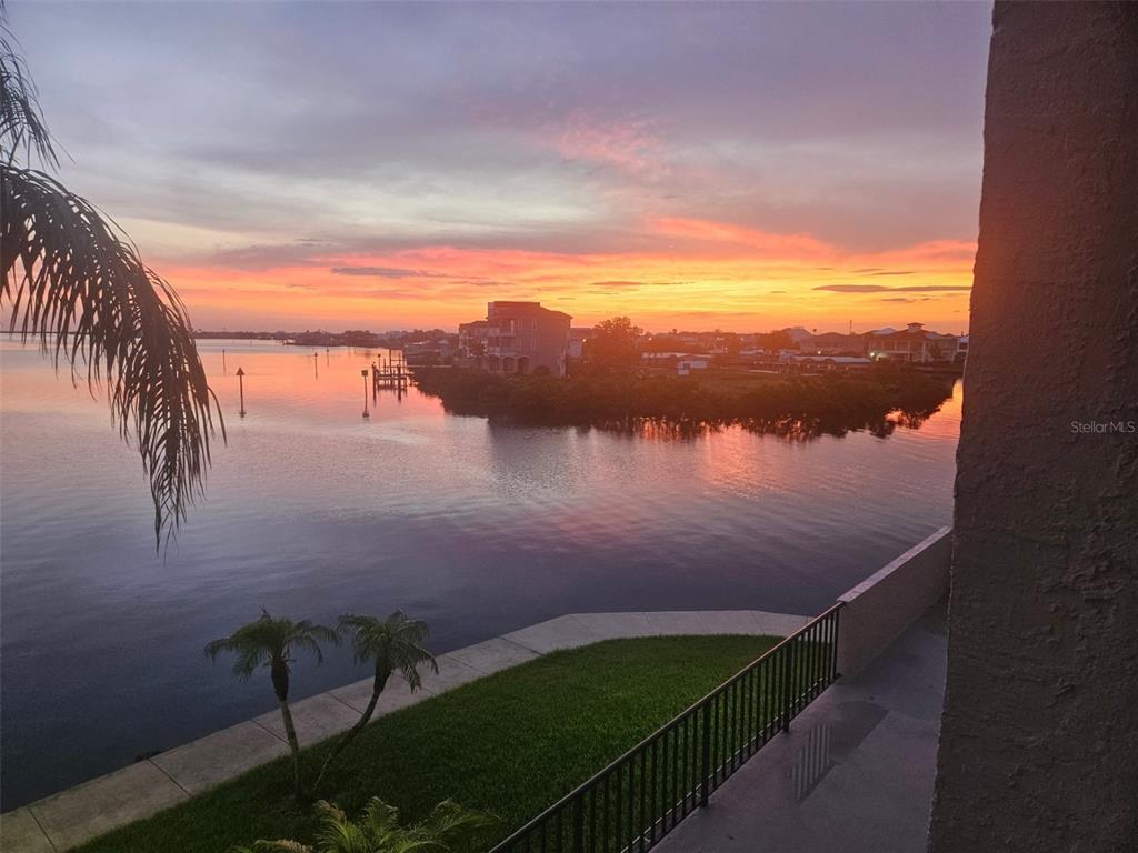 A balcony with blue chairs, a bench, and a view of the water and palm tree — 6035 SEA RANCH DRIVE UNIT 210, HUDSON, FL
