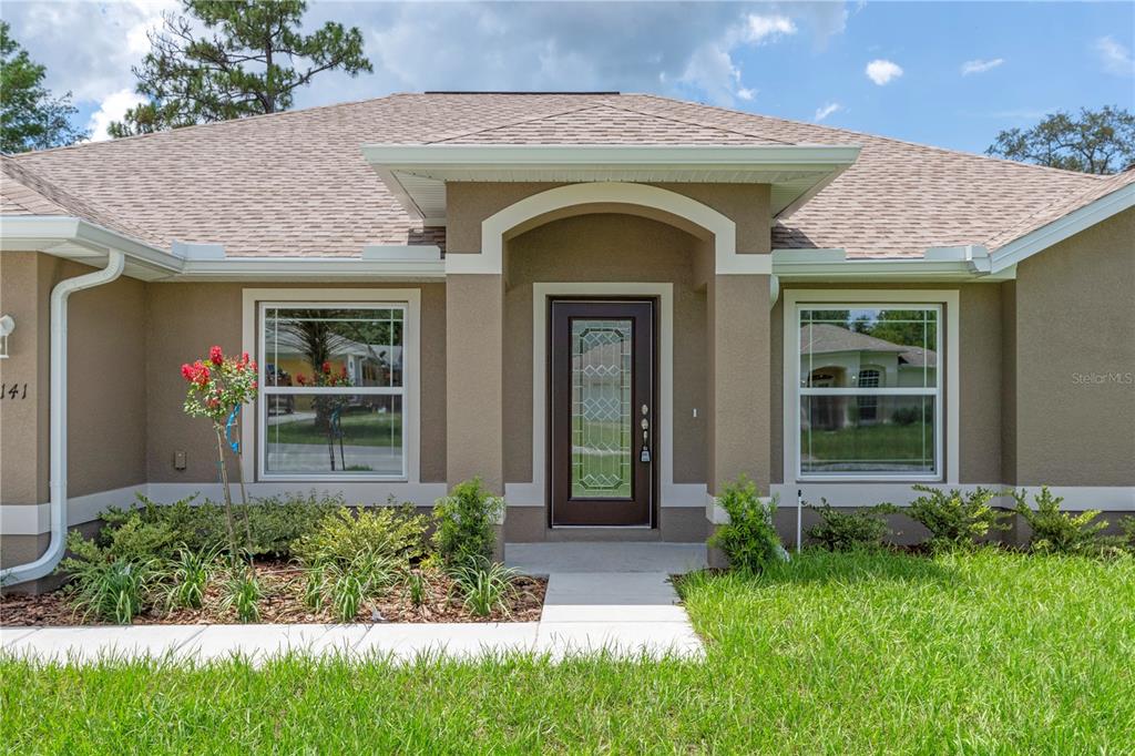 A single-story house exterior features a brown front door, tan stucco walls, and two windows — 13048 KITTIWAKE ROAD, WEEKI WACHEE, FL