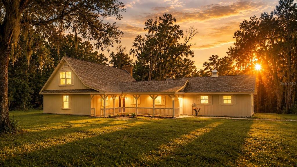 A light-colored farmhouse with a wrap-around porch sits on a grassy lawn at sunset — 3270 ENDSLEY ROAD, BROOKSVILLE, FL