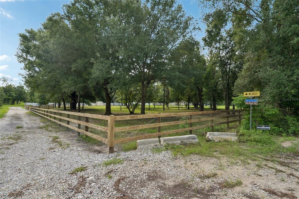 A fenced, grassy lot with mature trees sits beside a gravel road under a blue sky — 0 RAISER RD, DADE CITY, FL