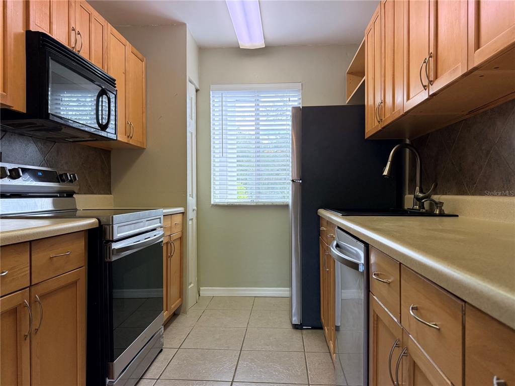 A galley kitchen features wood cabinets, stainless steel appliances, tile flooring, and a window — 6519 W NEWBERRY ROAD UNIT 305, GAINESVILLE, FL