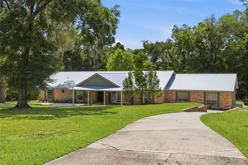 A single-story brick house with a metal roof sits behind a concrete driveway and lawn — 1150 MARKHAM WOODS ROAD, LONGWOOD, FL