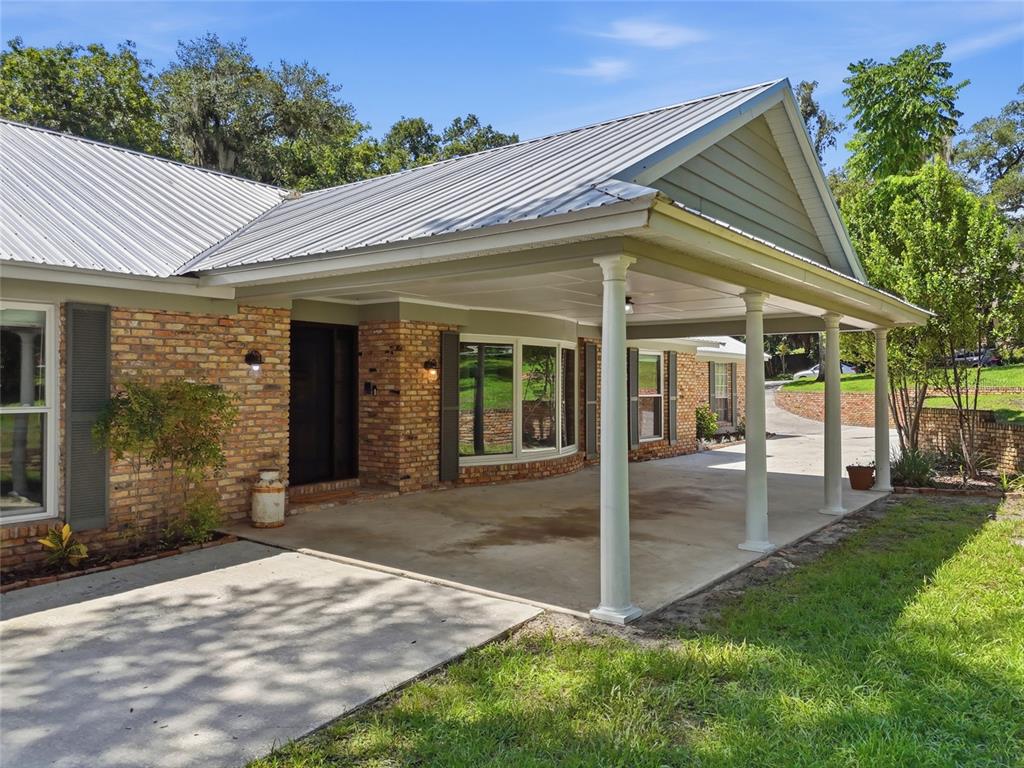 A brick house exterior features a metal roof, a covered carport, and white support columns — 1150 MARKHAM WOODS ROAD, LONGWOOD, FL