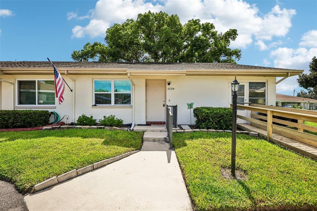 A single-story house with a white exterior, a concrete walkway, and a wooden wheelchair ramp — 2224 GRENADIER DRIVE, SUN CITY CENTER, FL
