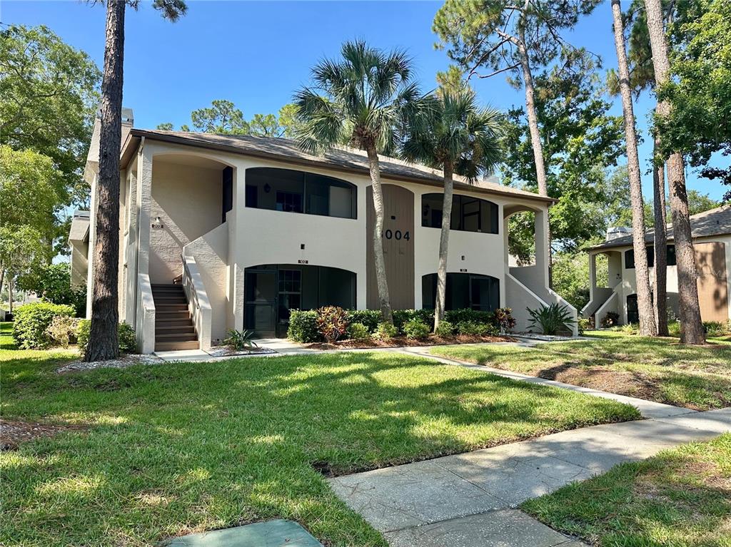 A two-story tan apartment building features screened balconies, exterior stairs, and surrounding palm trees — 3004 BONAVENTURE CIRCLE UNIT 102, PALM HARBOR, FL