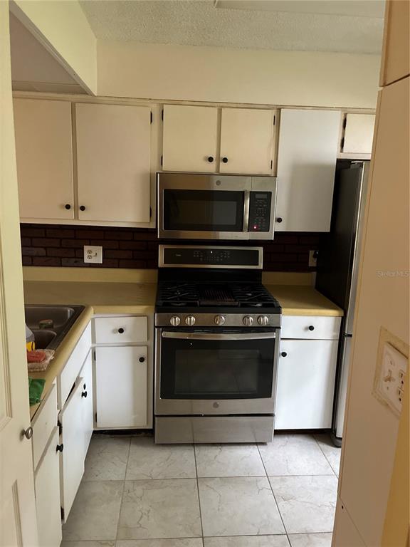 A kitchen features white cabinets, stainless steel appliances, a brick backsplash, and tiled flooring — 2968 FLINT DRIVE S, CLEARWATER, FL