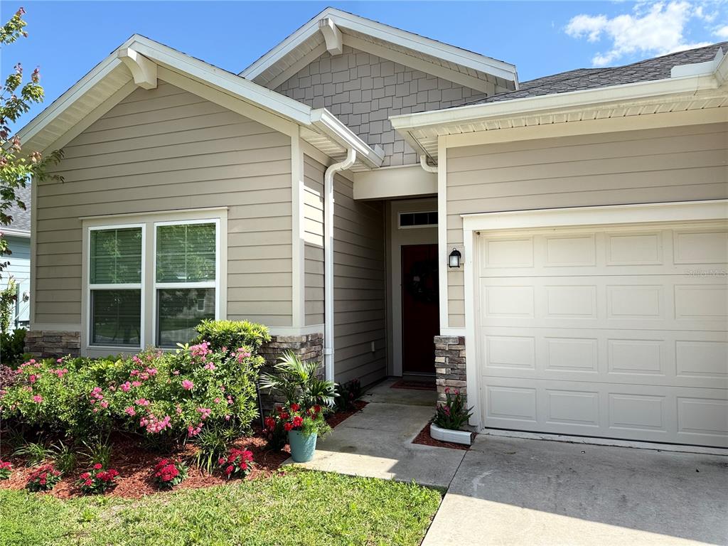 A tan house exterior features a front-facing garage, a recessed entryway, and landscaped flower beds — 7656 SW 74TH LOOP, OCALA, FL