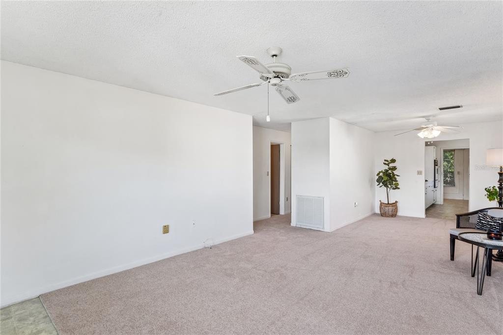 A carpeted living room with white walls, two ceiling fans, and a view into a kitchen — 5530 N 80TH STREET UNIT C308, ST PETERSBURG, FL