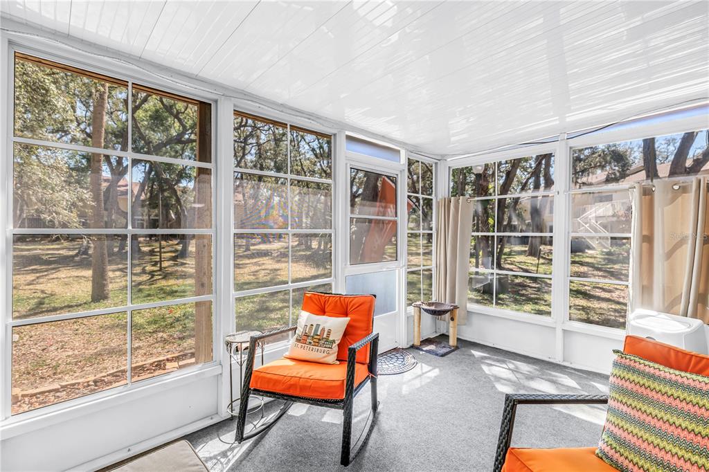 A sunroom with white-framed windows, gray carpeting, and orange patio furniture overlooking a wooded area — 3620 41ST WAY S UNIT F, ST PETERSBURG, FL