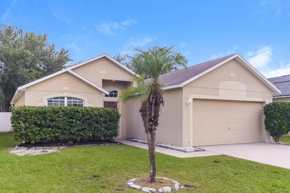 A single-story tan house with a two-car garage, a palm tree, and a manicured hedge — 5470 SILVER THISTLE LANE, ST CLOUD, FL