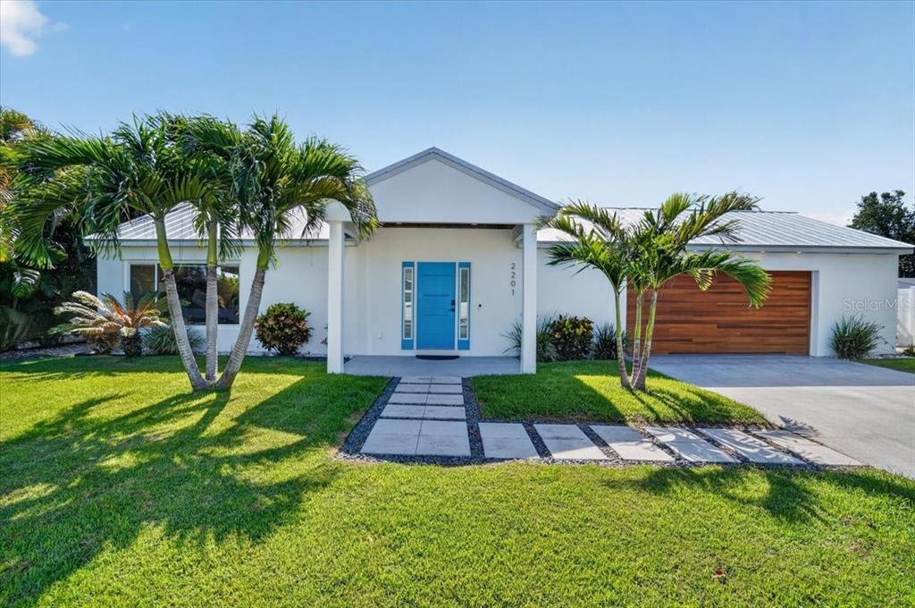 A single-story white house features a bright blue front door and a wood-paneled garage door — 2201 WEBBER STREET, SARASOTA, FL