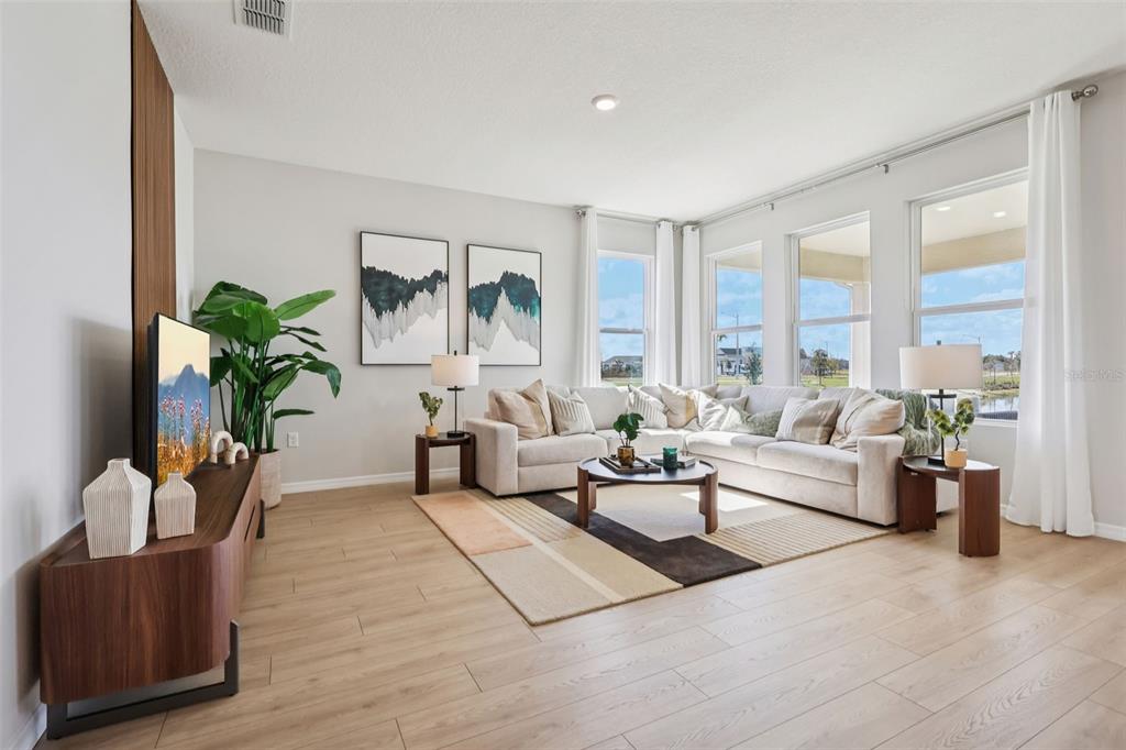 A bright living room features a sectional sofa, wood-look flooring, and large windows with curtains — 8595 HEMPSTEAD WAY, MOUNT DORA, FL