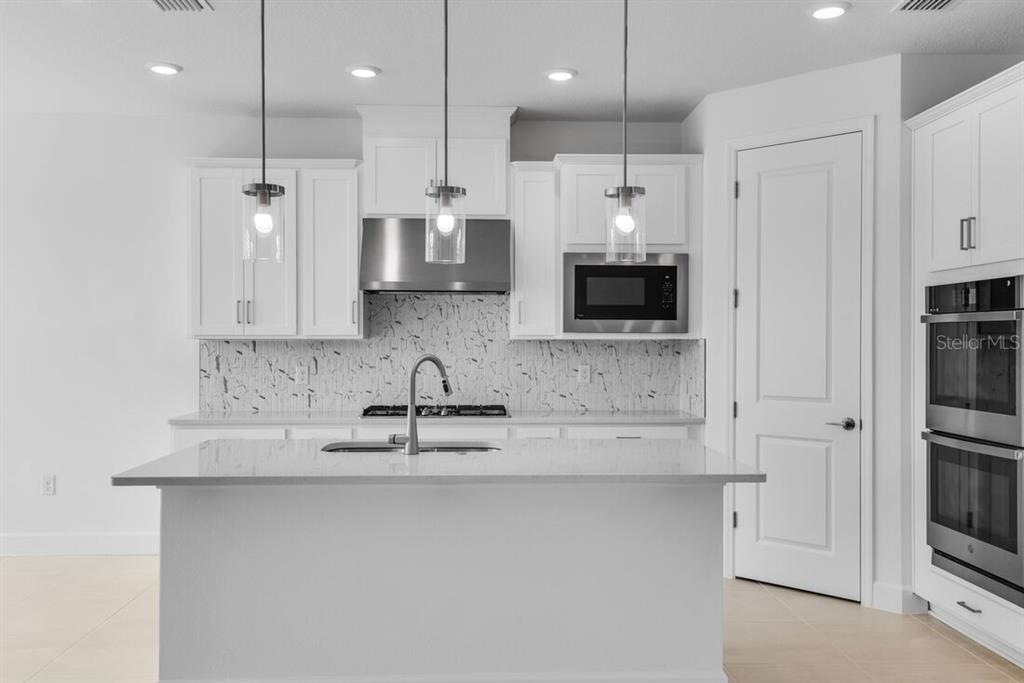 A modern white kitchen features a central island, stainless steel appliances, and marble-patterned backsplash tile — 672 JUNONIA BOULEVARD, NEW SMYRNA BEACH, FL