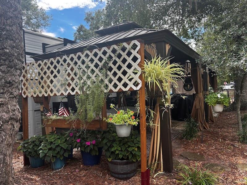 A metal-roofed gazebo features a white lattice screen, hanging plants, and potted flowers — 14105 253RD AVENUE ROAD NE UNIT 392, FORT MC COY, FL