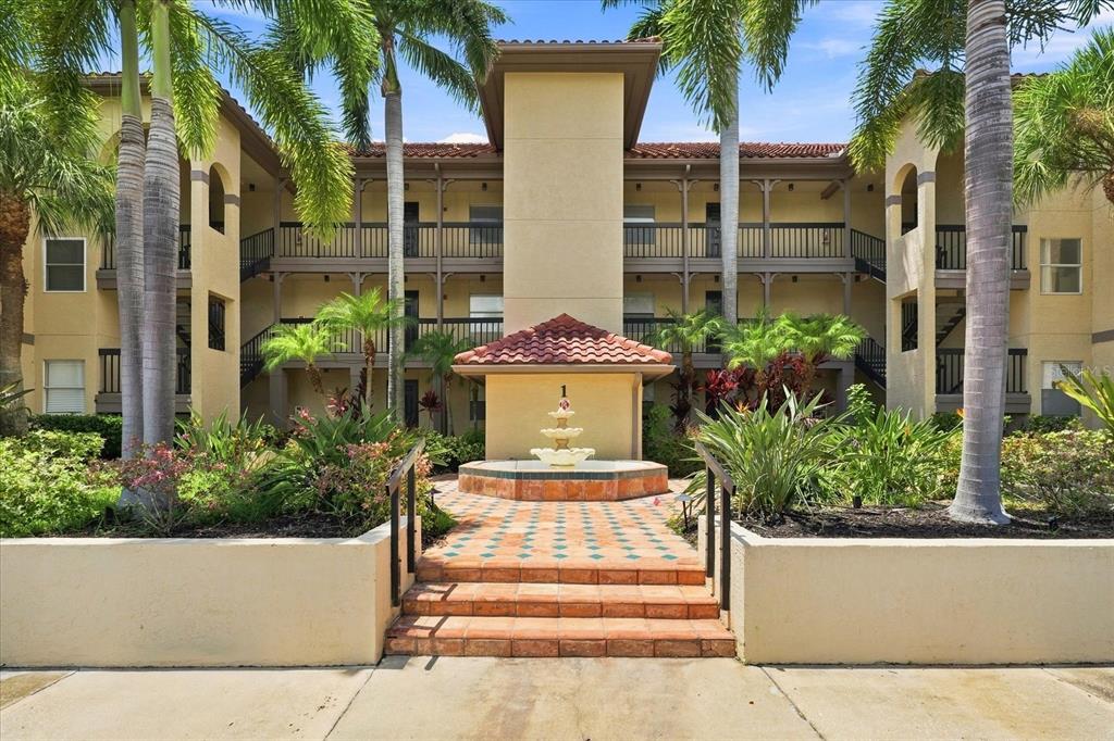 A three-story apartment building features a central fountain, tiled steps, and surrounding palm trees — 2400 FEATHER SOUND DRIVE UNIT 133, CLEARWATER, FL