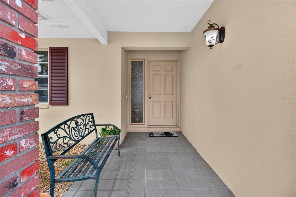 A covered entryway features a tiled floor, a metal bench, and a tan front door — 2221 COLONIAL DRIVE, DUNEDIN, FL