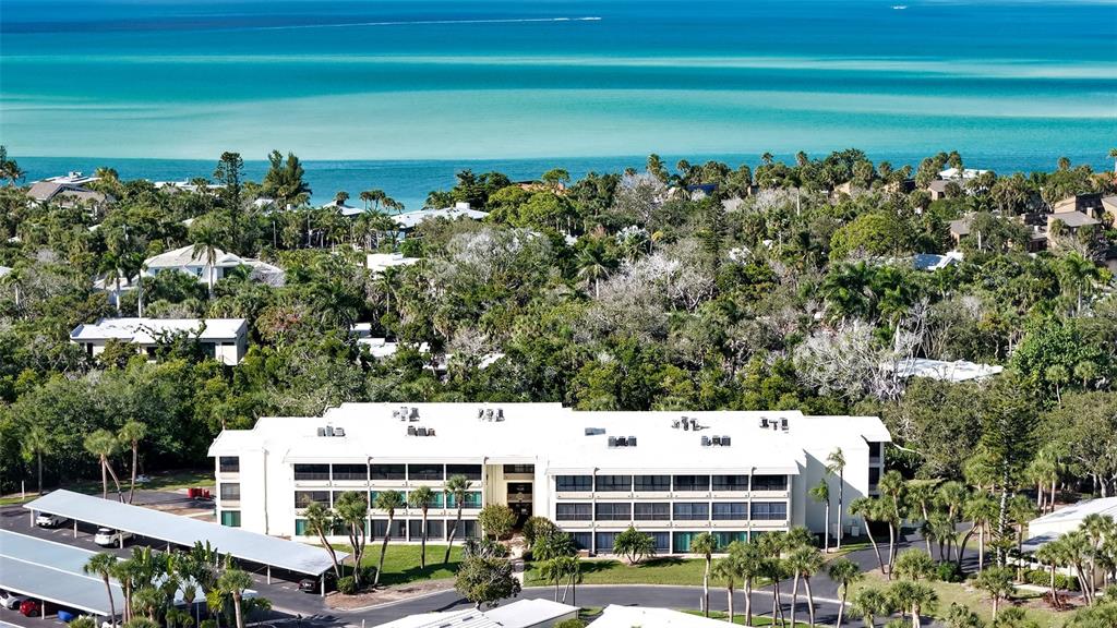 An aerial view shows a white multi-story apartment building surrounded by lush trees near water — 225 HOURGLASS WAY UNIT 103, SARASOTA, FL