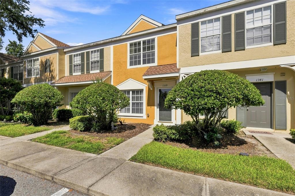 A two-story townhouse exterior features yellow stucco walls, dark shutters, and small front yard landscaping — 1339 COSTA MESA DRIVE, WESLEY CHAPEL, FL