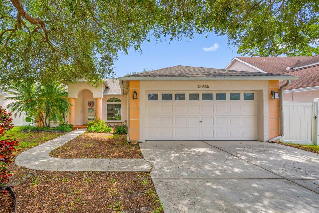 A single-story house with a two-car garage, light-colored stucco walls, and a concrete driveway — 12965 PINEFOREST WAY W, LARGO, FL