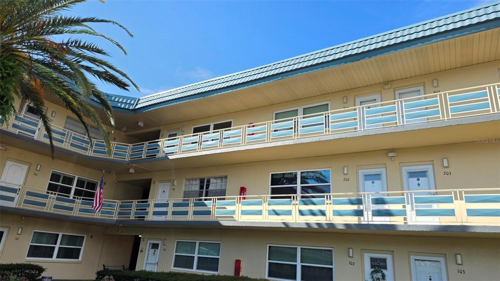 A three-story apartment building exterior features tan walls, blue railings, and numbered unit doors — 5750 80TH STREET N UNIT C202, ST PETERSBURG, FL