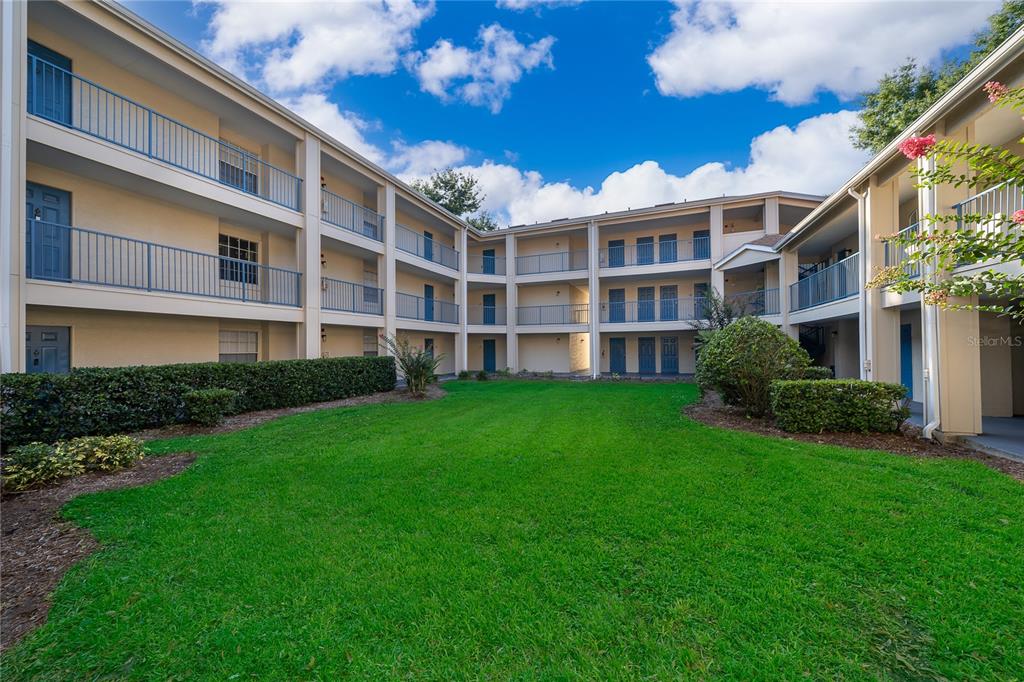 A three-story apartment building surrounds a central grassy courtyard under a blue sky — 7210 WESTPOINTE BOULEVARD UNIT 1312, ORLANDO, FL