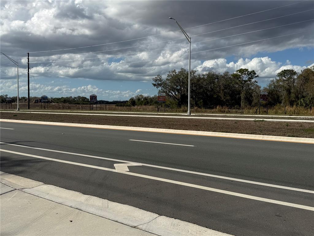 A paved road runs alongside a grassy median and an undeveloped field under a cloudy sky — 11959 MOCCASIN WALLOW ROAD, PARRISH, FL