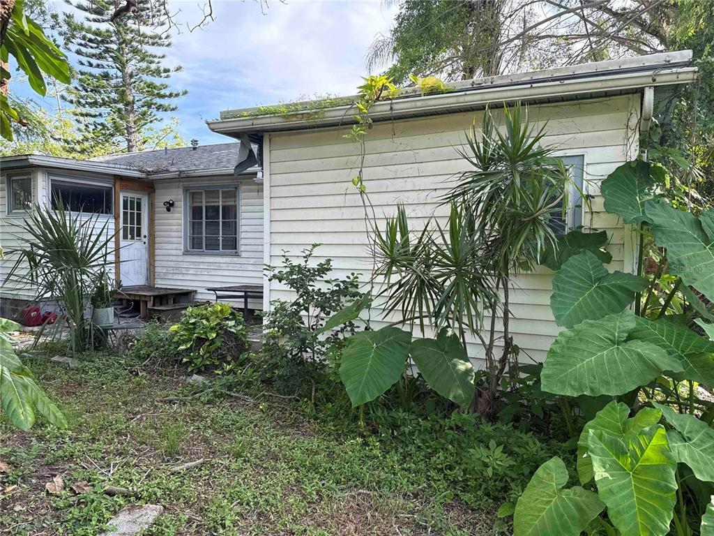 The exterior of a white-sided house features overgrown vegetation and a small wooden porch — 7011 HIBISCUS AVENUE S, SOUTH PASADENA, FL
