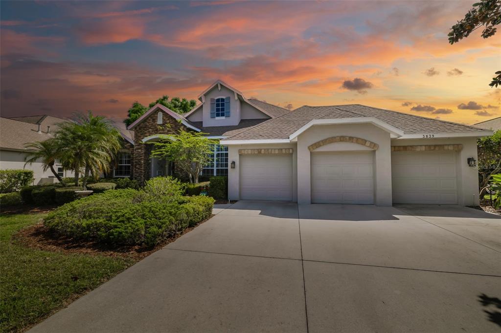 A single-story house with a three-car garage, stone accents, and a concrete driveway at sunset — 3839 59TH AVENUE CIRCLE E, ELLENTON, FL
