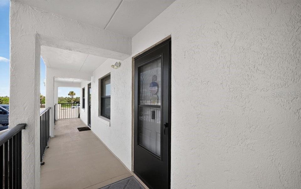 A covered exterior walkway leads to a black front door on a white stucco building — 2700 OCEAN SHORE BOULEVARD UNIT 208, ORMOND BEACH, FL