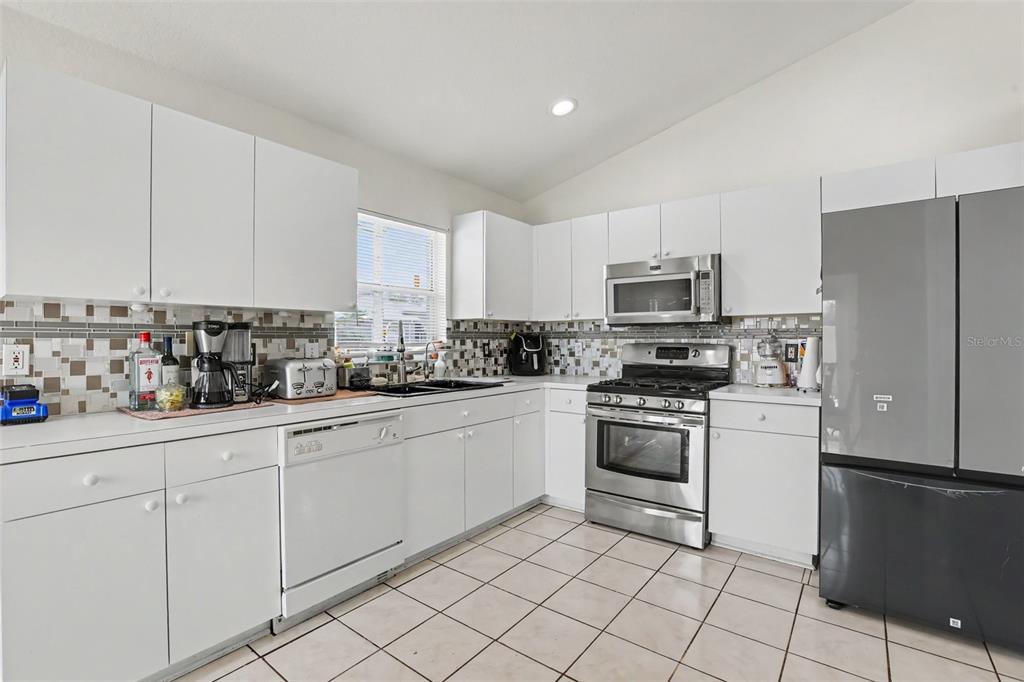 A kitchen features white cabinets, stainless steel appliances, a tiled backsplash, and light-colored floor tiles — 1102 VINETREE DRIVE, BRANDON, FL