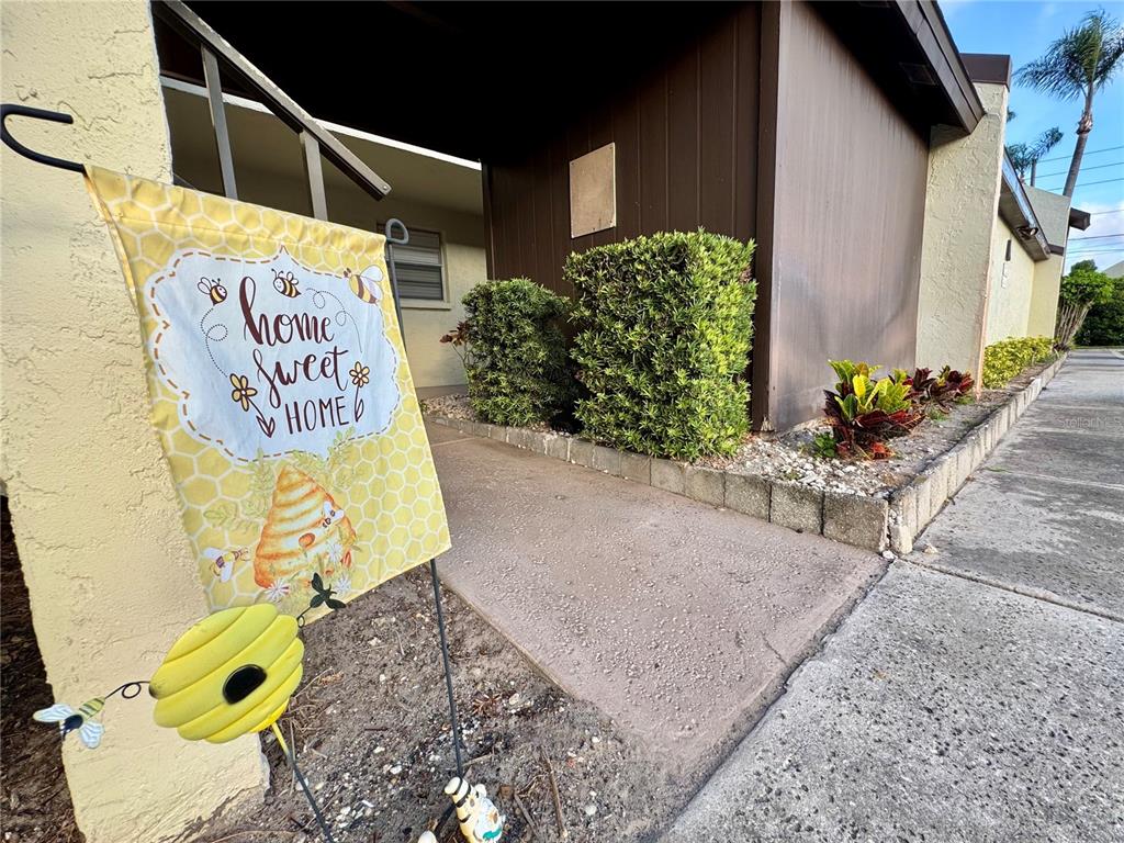 A yellow garden flag stands on a walkway leading to a building entrance with hedges — 1100 E BAY DRIVE UNIT 3, LARGO, FL