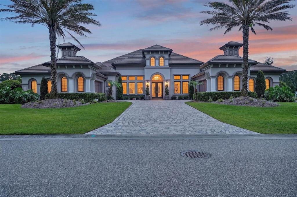 A symmetrical luxury house with a paved driveway, palm trees, and arched windows at dusk — 1023 SUDBURY LANE, ORMOND BEACH, FL