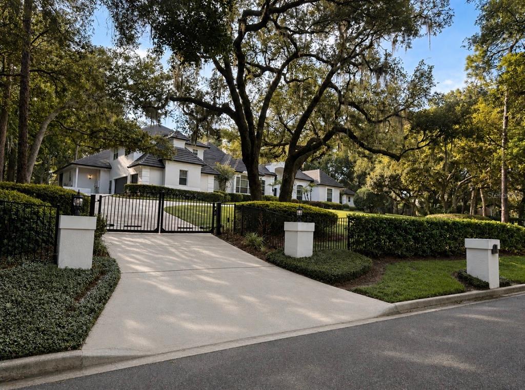 A gated concrete driveway leads to a white multi-story house surrounded by large oak trees — 950 SWEETWATER CLUB BOULEVARD, LONGWOOD, FL