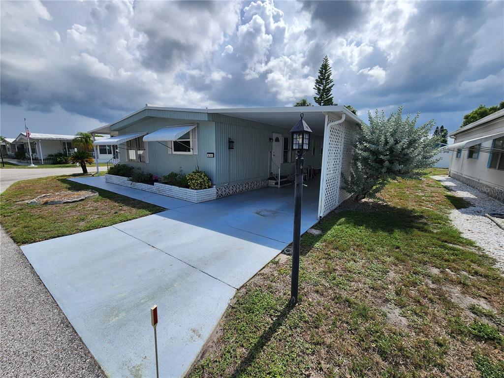 A light green mobile home features a carport, a concrete driveway, and a small garden — 808 53RD AVENUE E UNIT 223, BRADENTON, FL