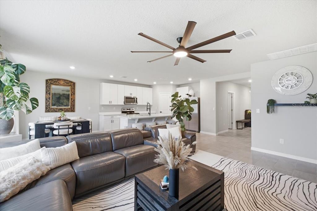 An open-concept living room and kitchen featuring a dark leather sectional, ceiling fan, and dining area — 10119 DAYBREAK GLEN, PARRISH, FL
