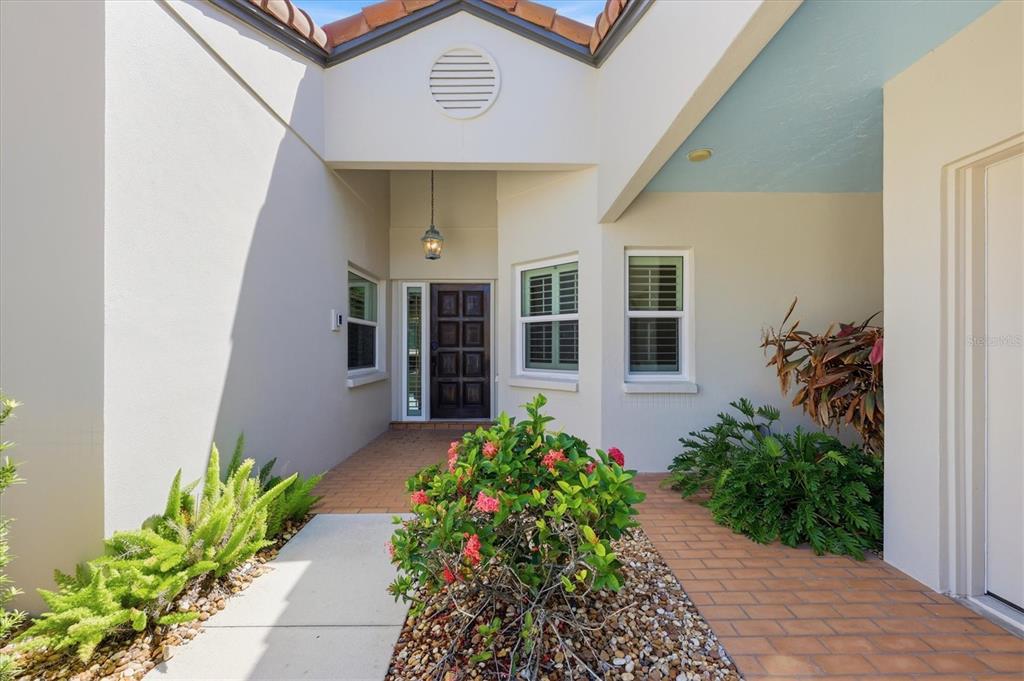 The exterior entryway features a dark wood front door, brick walkway, and landscaped garden beds — 7765 CALLE FACIL, SARASOTA, FL