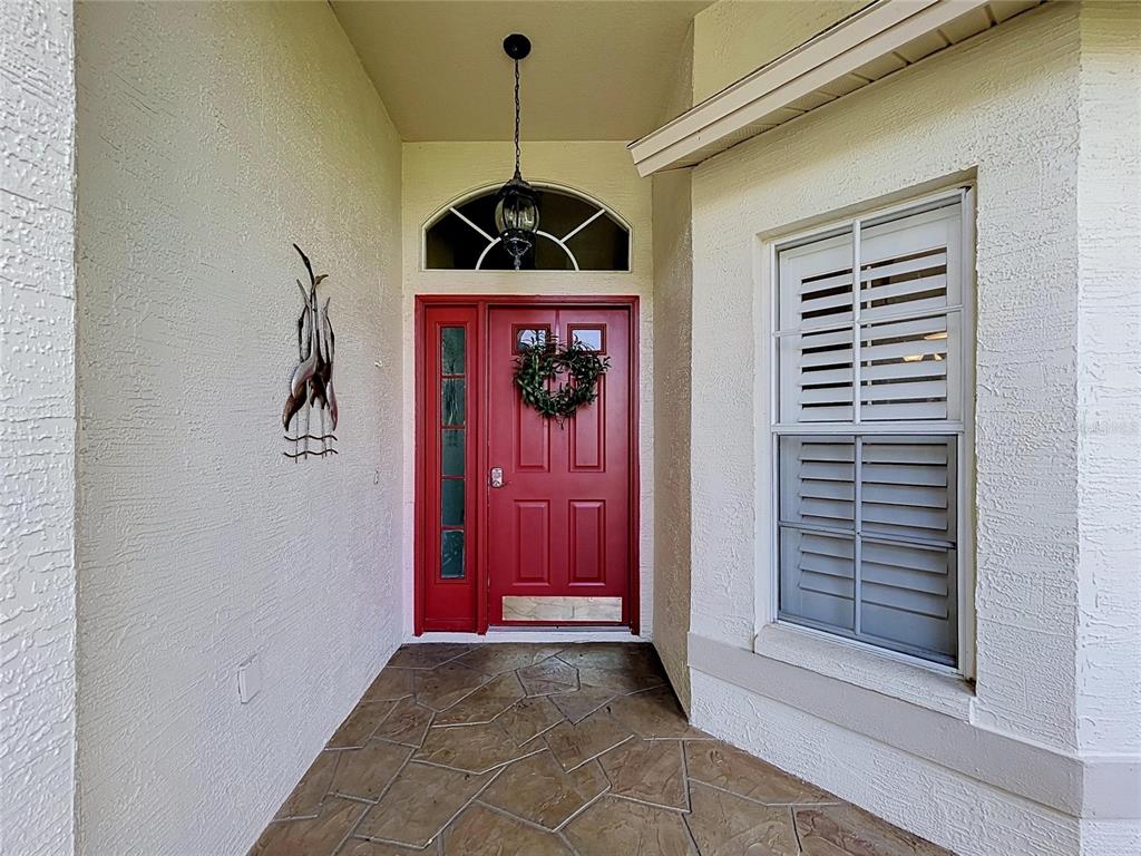 A red front door with a sidelight and wreath sits under a covered entryway — 1017 SWEET JASMINE DRIVE, TRINITY, FL